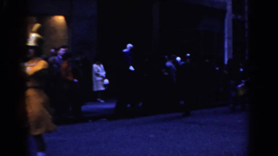 A blurred yellow-uniformed marcher moves out of frame as spectators remain near a building entrance in 1962 New York City footage.