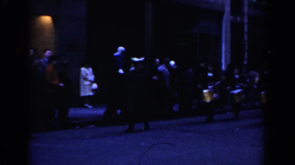 Nighttime 1962 New York City street frame with spectators gathered by a building entrance after the foreground performer has passed.