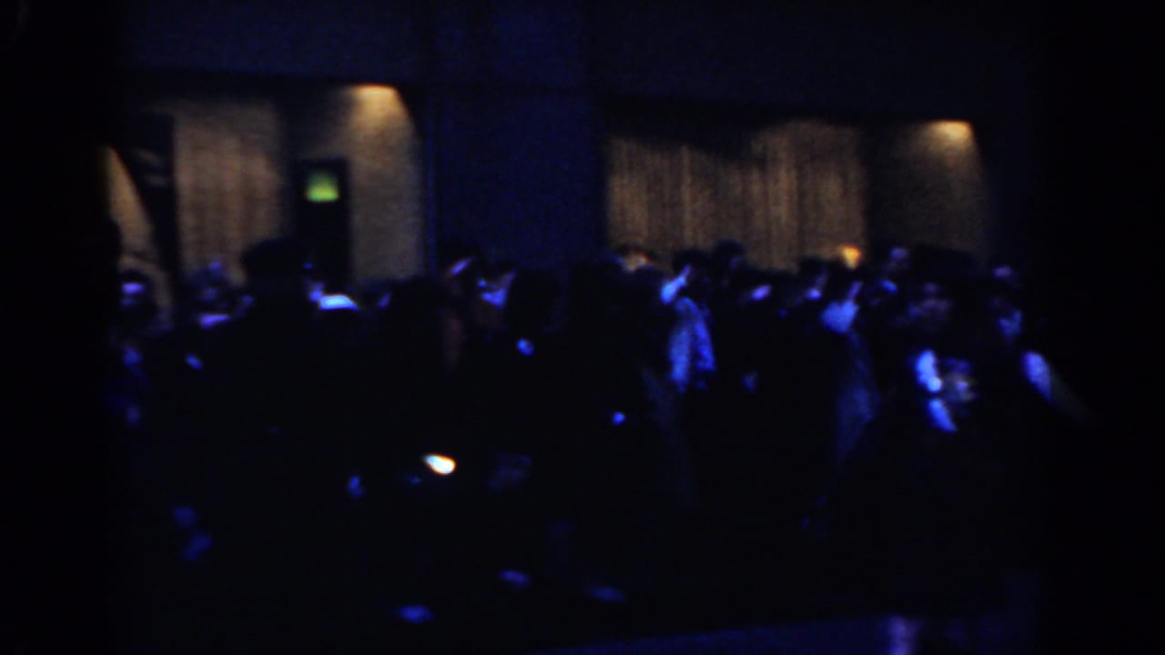 Nighttime parade crowd with kilted band members under blue lighting.