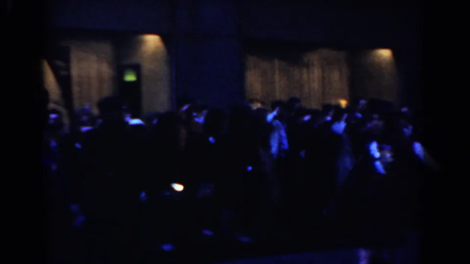 1962 New York City home-movie frame showing a nighttime parade crowd and kilted marching band in dim blue street light.