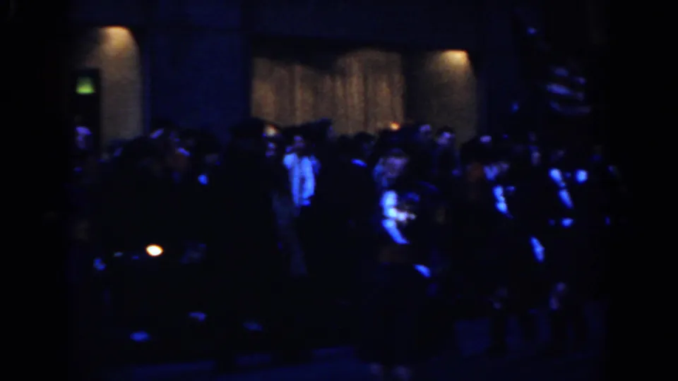 Nighttime New York City parade frame with spectators and marching performers visible in low blue light.