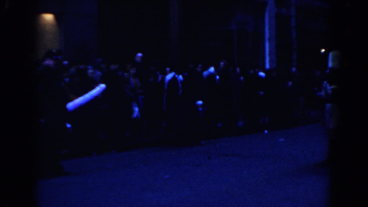Blue-lit nighttime street crowd with a marching group moving past spectators.