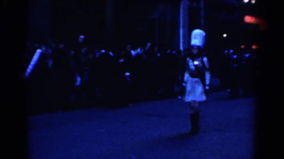 1962 New York City frame with a woman in a white dress and tall white hat leading or standing before a dark marching group.
