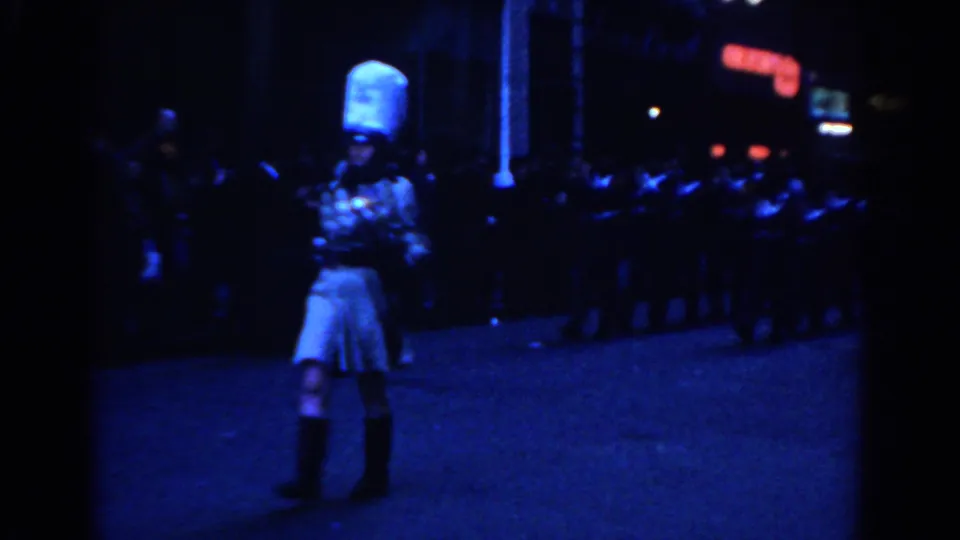1962 New York City 8mm frame showing a white-costumed woman with tall hat before a row of marchers under blue night lighting.