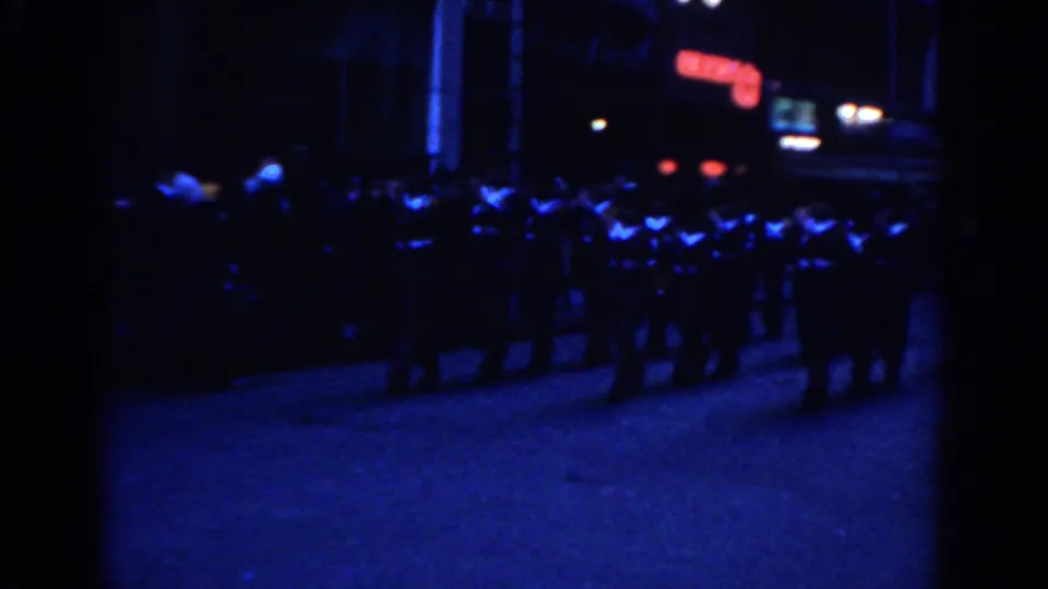 1962 New York City frame showing a row of marchers crossing a blue-lit street with a red neon sign in the distance.