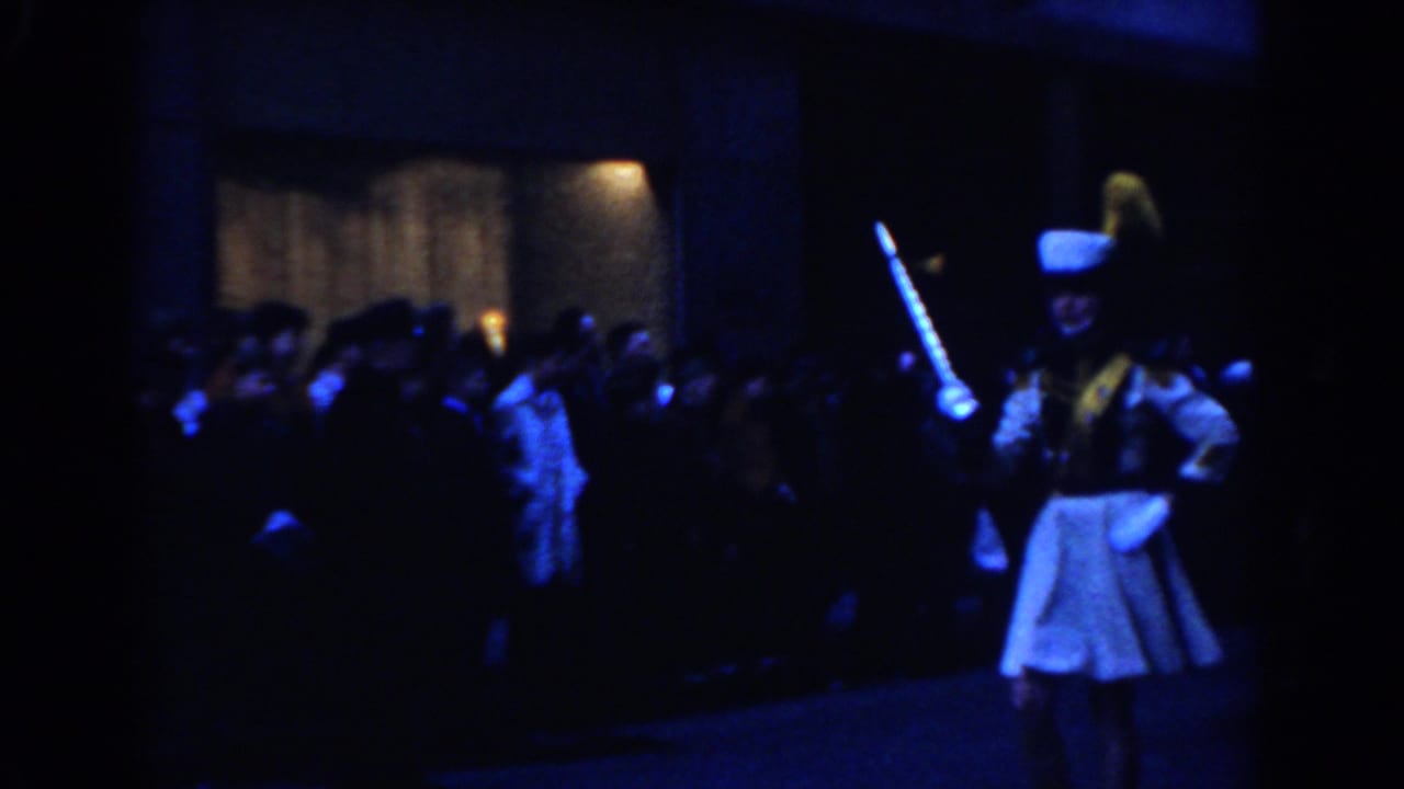 Majorette in white uniform marching at night while spectators watch from the sidewalk.