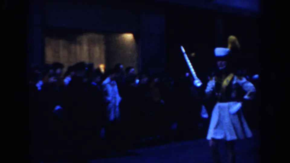 1962 New York City home-movie frame showing a majorette or parade leader in a white uniform and tall hat passing a nighttime crowd.