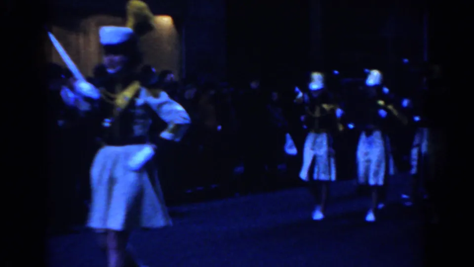 A white-uniformed parade leader with a baton marches through a dark New York City street, with spectators and band members behind.