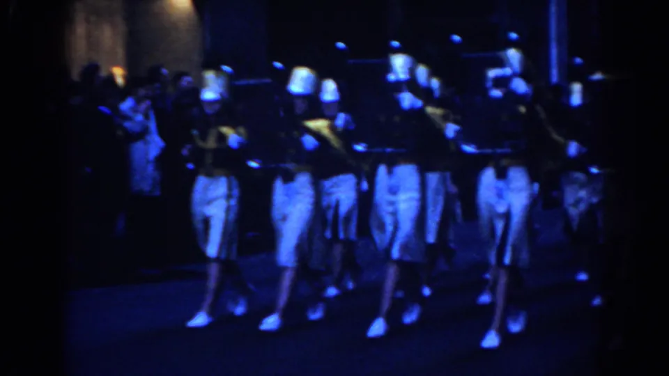1962 New York City parade footage shows marching band members in white uniforms and tall hats advancing toward the camera at night.
