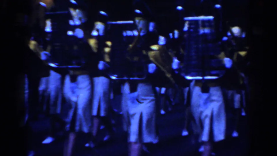 Marching musicians in tall hats and white trousers fill the frame during a nighttime New York City parade captured on 8mm film.