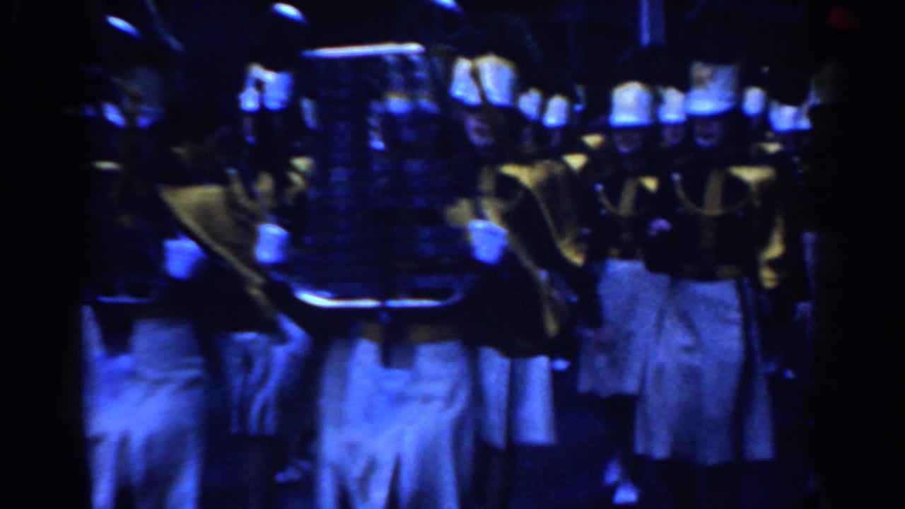 Marching band members in white caps and gold-toned jackets move through a dark street.