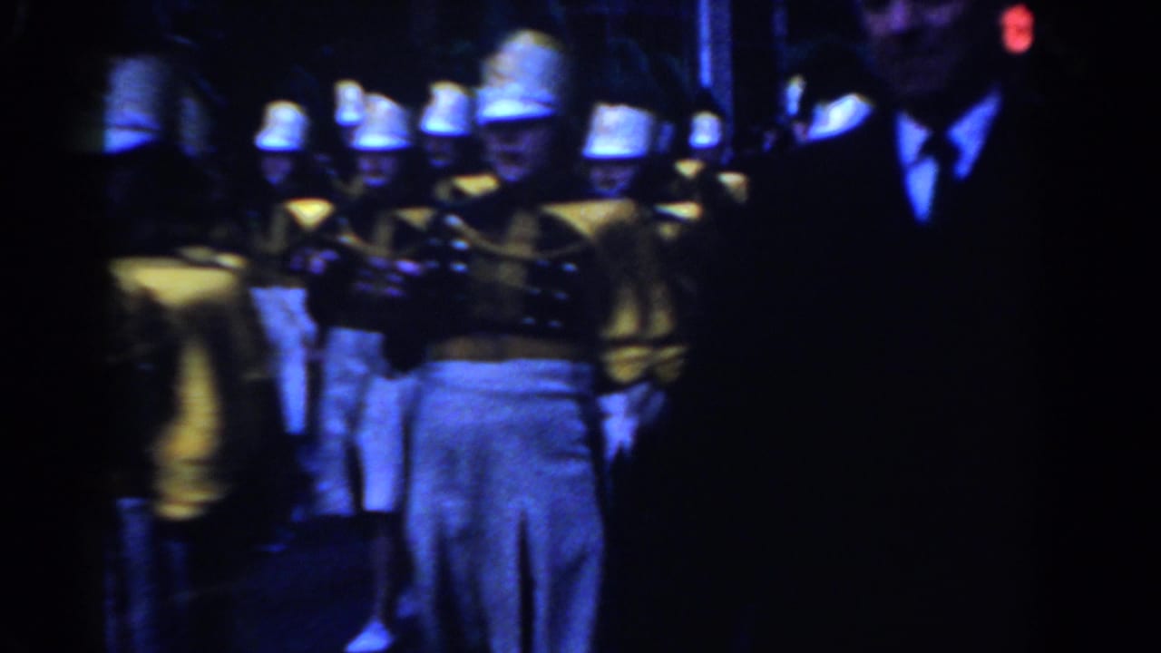 Uniformed performers march in formation while a suited spectator stands at the edge of the frame.