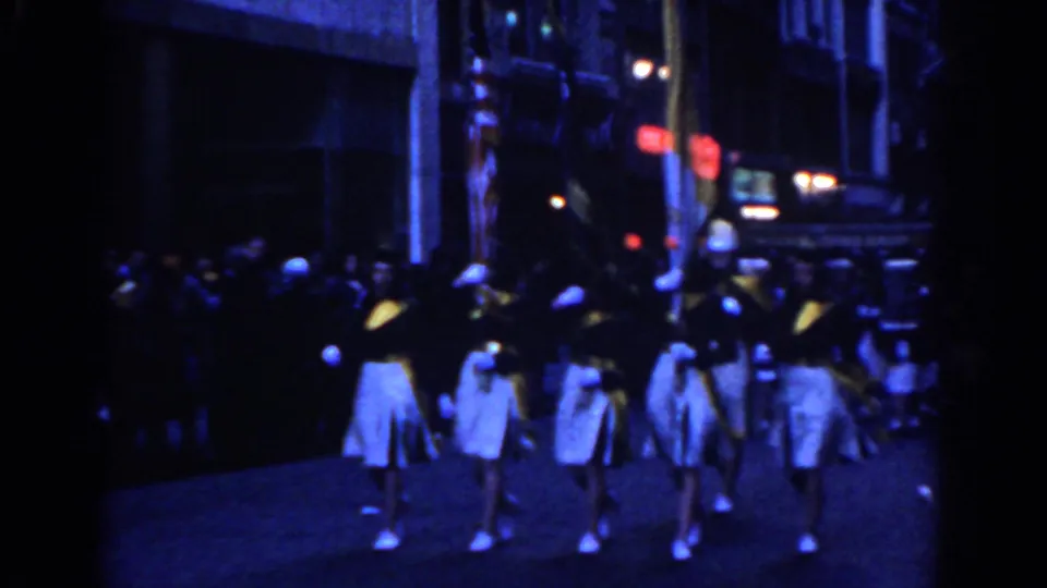 Wide 1962 New York City 8mm view of a marching band proceeding along a nighttime street with spectators nearby.