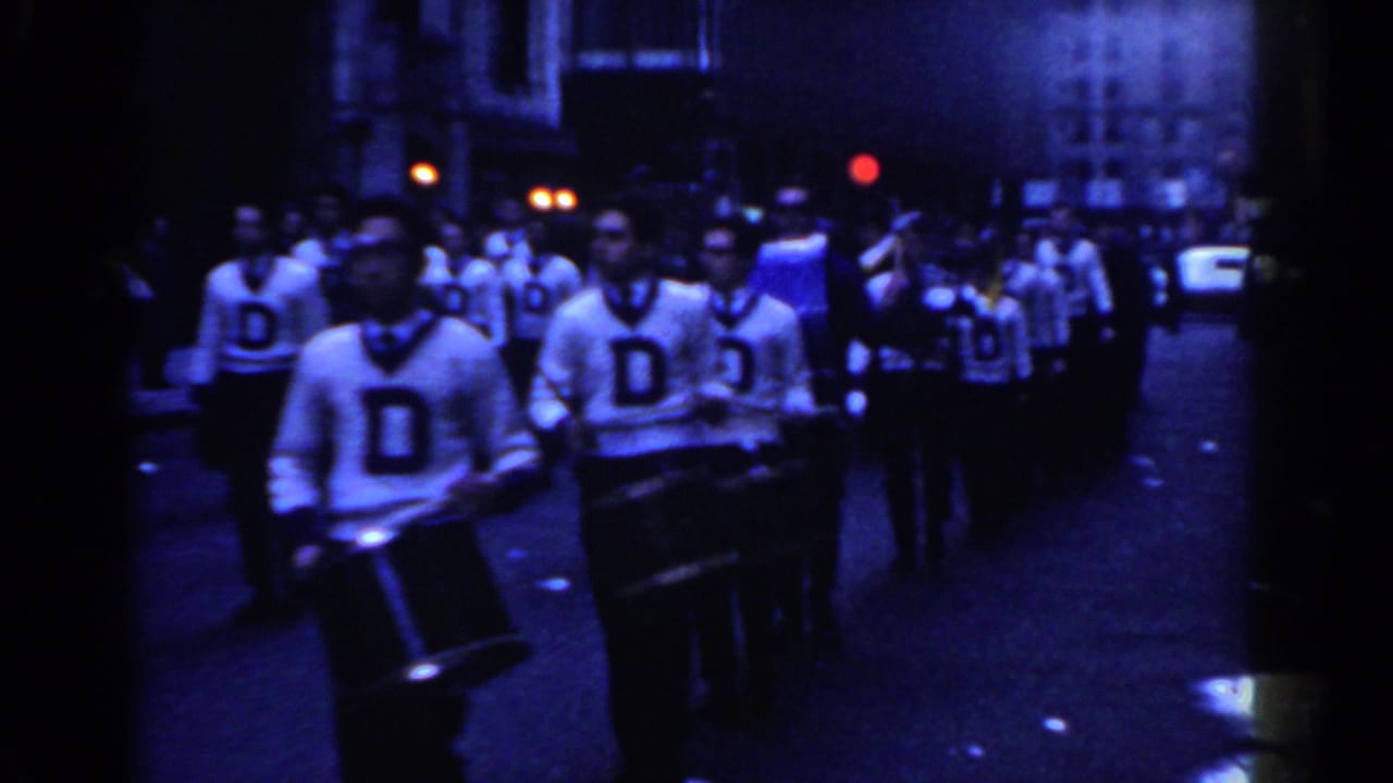 1962 New York City home-movie frame showing student drummers in white sweaters with a large letter marching at night.