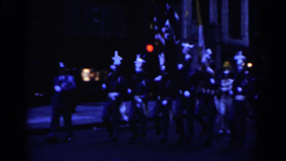 Nighttime parade frame with flag bearers and a color guard moving through a New York City street in 1962.