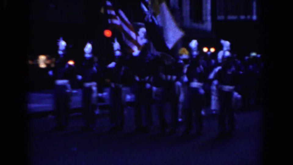 1962 New York City parade frame showing flags, color guard members, and drummers in low-light 8mm footage.