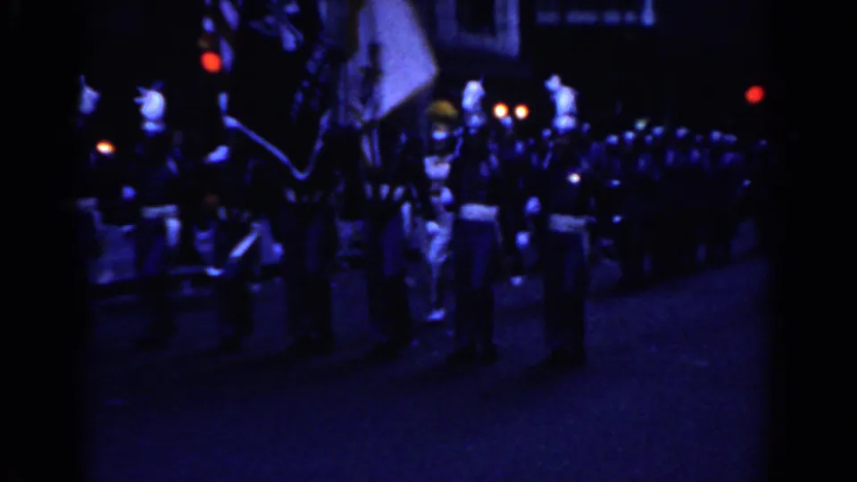 Nighttime 1962 New York City parade frame with a flag line and uniformed marchers continuing down the street.