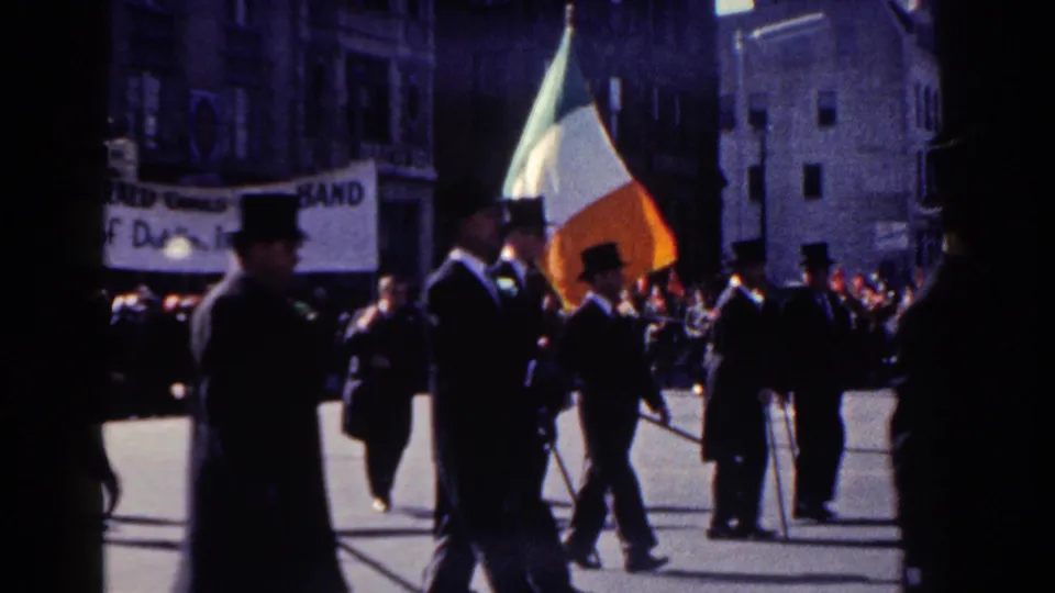 Men in formal dark suits and hats pass an Irish tricolor flag and pipe band banner in New York City parade footage.