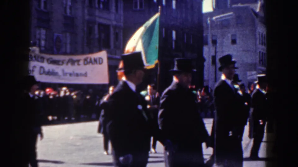 Suited marchers in hats cross the frame while a banner reading as a pipe band of Dublin appears behind them.