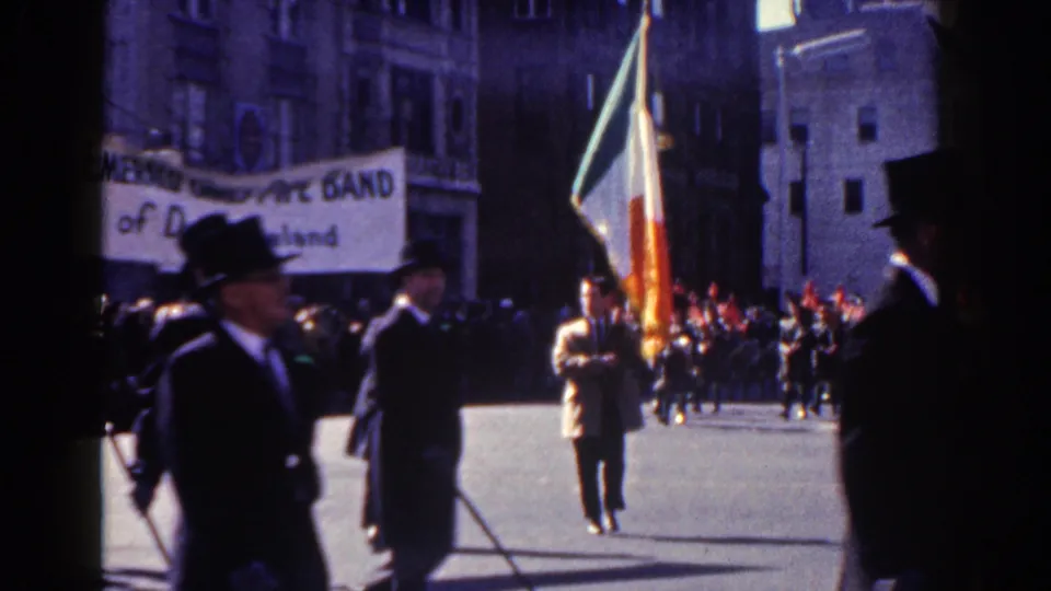 An Irish tricolor flag bearer and formal marchers move along the New York City parade route with spectators in the background.