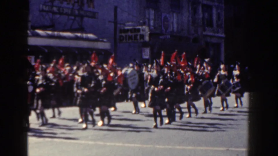 1962 New York City parade frame showing a pipe band in kilts with red plumes marching past spectators and visible diner signage.