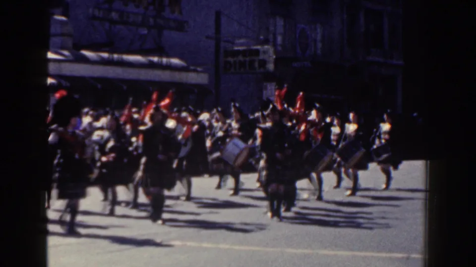 1962 New York City street parade with kilted musicians, red-plumed hats, drums, and bagpipes moving past period storefront signs.