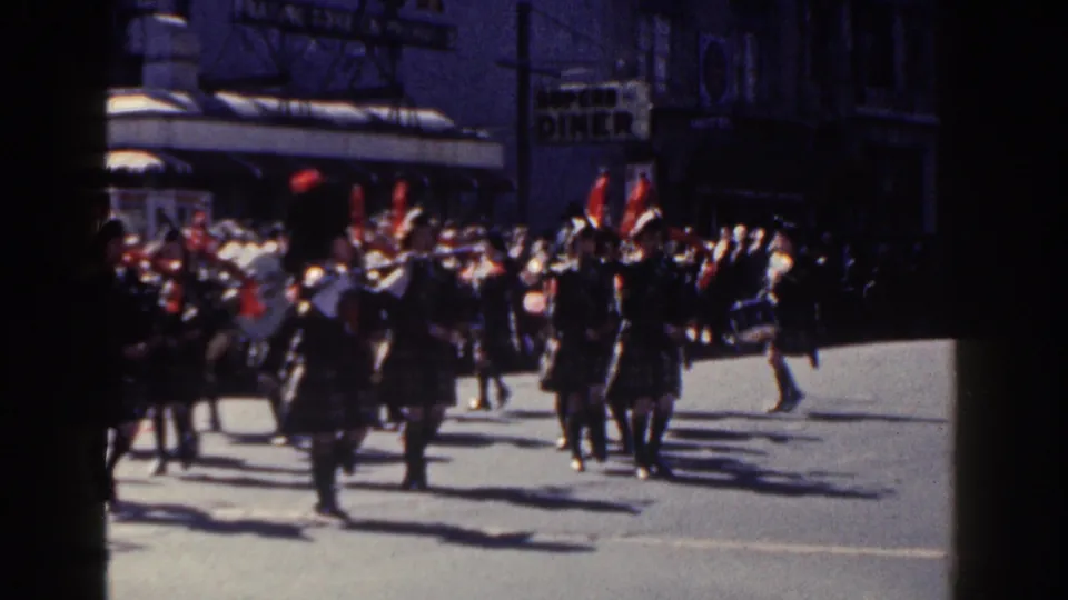 Final 1962 New York City parade frame with pipe band members in formation, red plumes, instruments, spectators, and storefront signage.