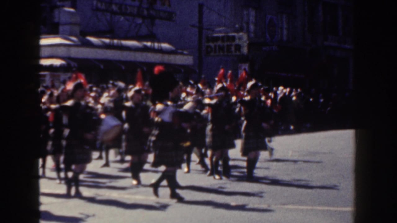 In 1962 New York City home-movie footage, a female marching band in tartan uniforms moves down a sunlit street with brass instruments, drums, spectators, and storefront signage behind them.