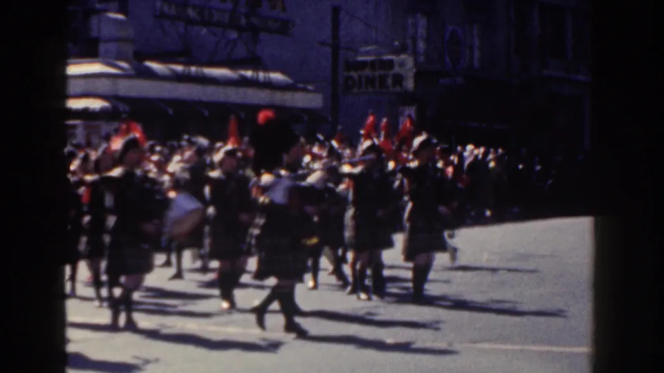 In 1962 New York City home-movie footage, a female marching band in tartan uniforms moves down a sunlit street with brass instruments, drums, spectators, and storefront signage behind them.