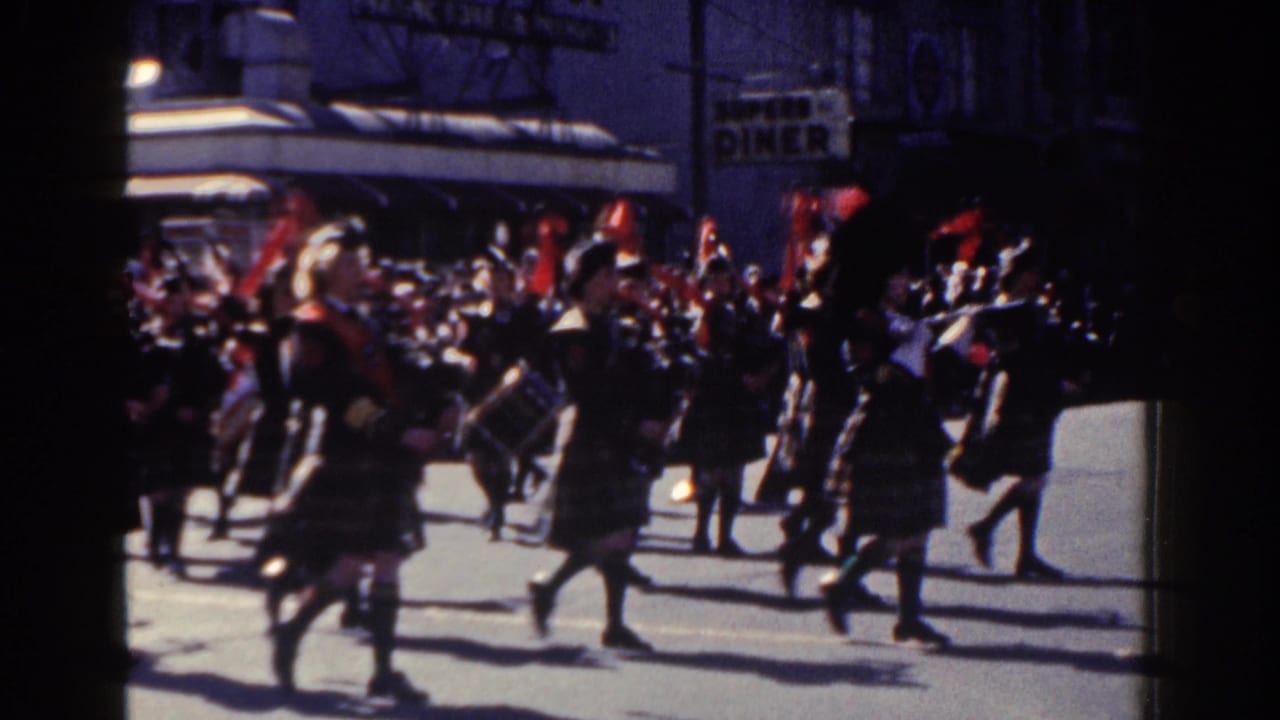Band members in tartan uniforms march with drums and brass instruments past a diner sign.