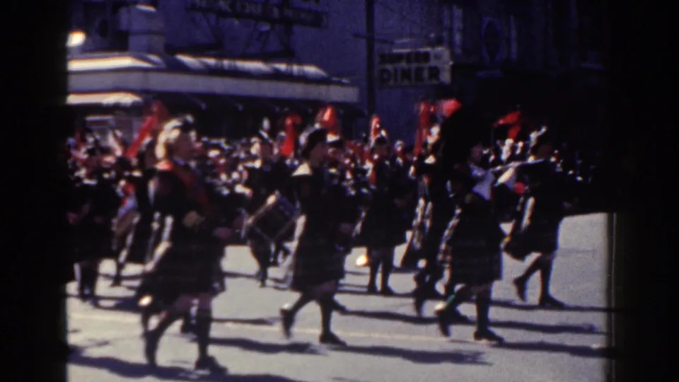 Female musicians in tartan kilts and dark jackets march through a 1962 New York City parade, with drums, brass instruments, red plumes, spectators, and a visible diner sign in the background.