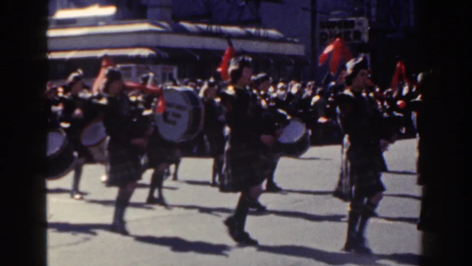 A handheld 8mm frame from the 1962 New York City parade shows drummers and brass players in tartan uniforms moving across the street, with spectators and commercial signage partly visible behind them.