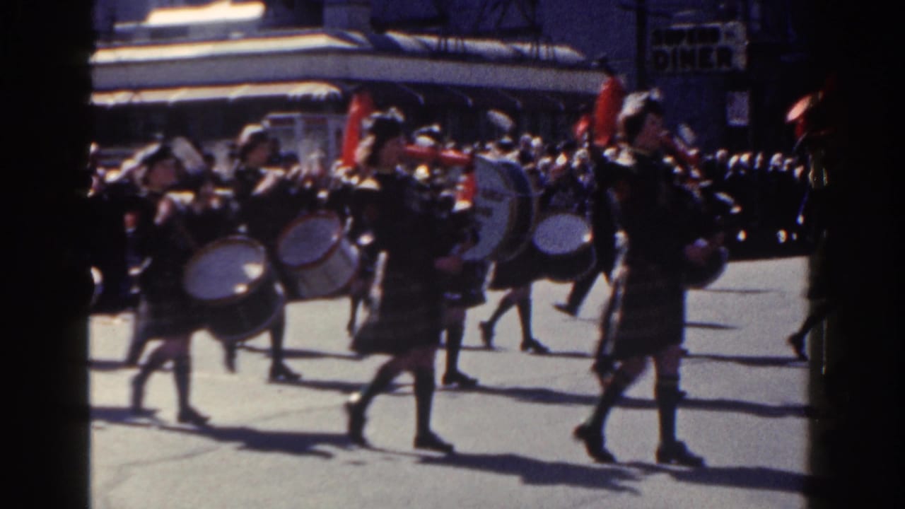 Kilted drummers march across a city street during a parade.