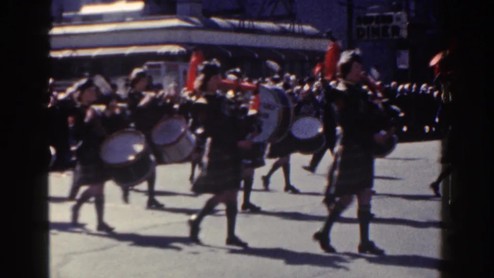 1962 New York City home-movie frame showing kilted drummers marching past spectators on a sunlit street.