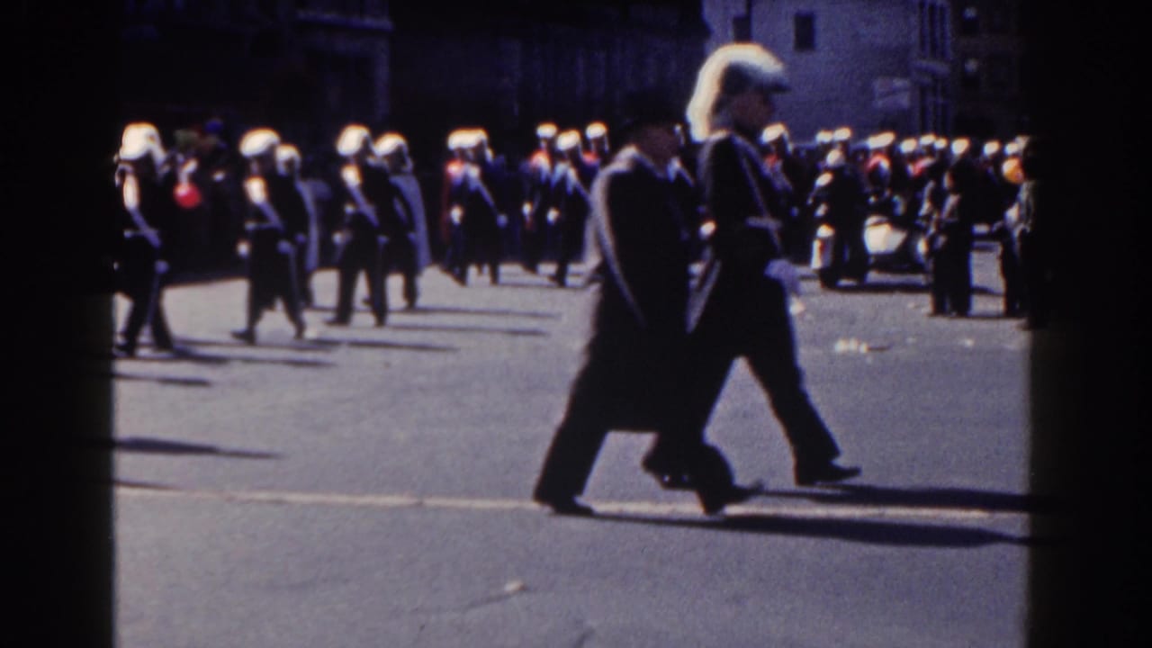 Uniformed marchers with white plumed hats cross a broad street.