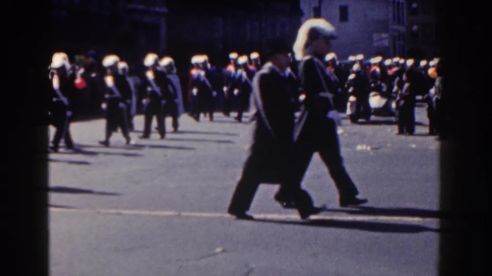 Uniformed parade unit with white plumed hats marches through a New York City street in the 1962 home movie.