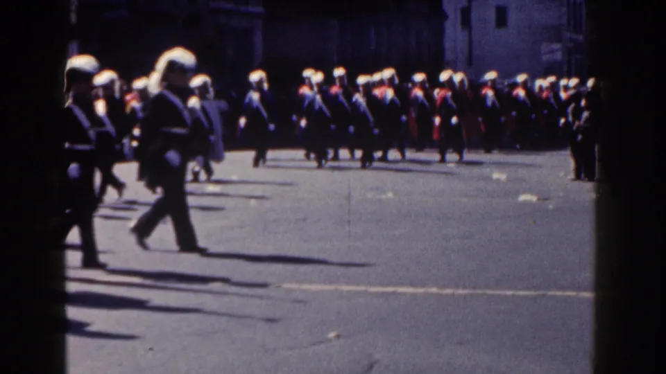 1962 New York City parade frame with uniformed marchers in plumed hats moving past curbside spectators.