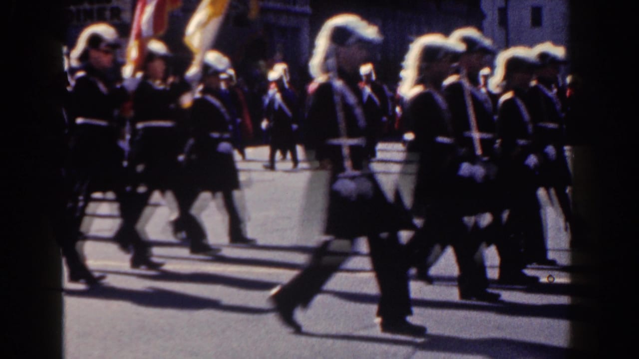 Uniformed marchers with white plumes pass along a city street during a parade.