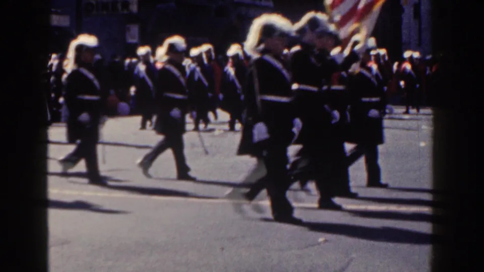 Uniformed marchers in plumed hats move across a New York City parade route, with pedestrians and storefronts in the background.