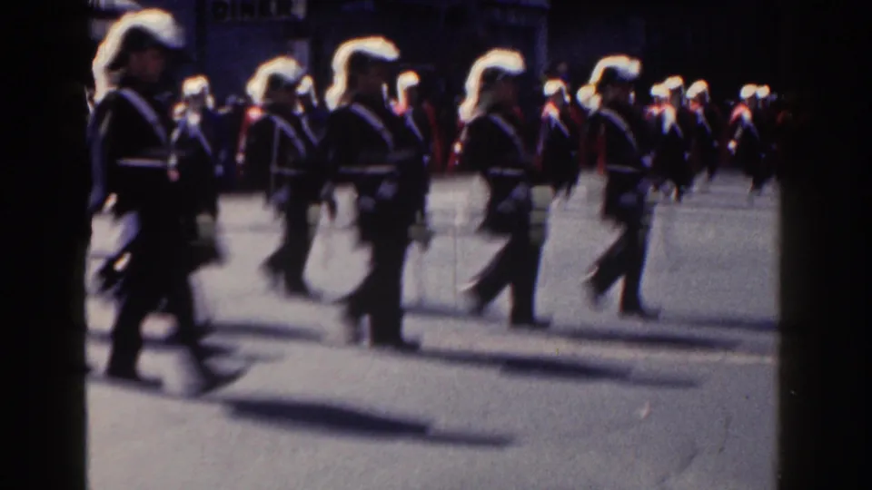 A soft, motion-blurred 8mm frame shows uniformed parade marchers passing close to the camera on a New York City street.