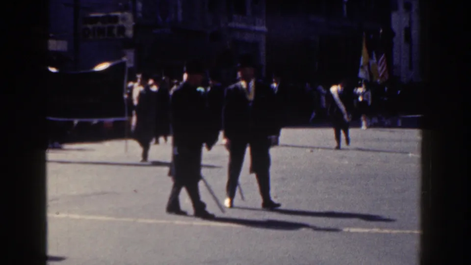Wider 1962 New York City parade view with uniformed figures, American flags, pedestrians, and a visible diner sign in the background.