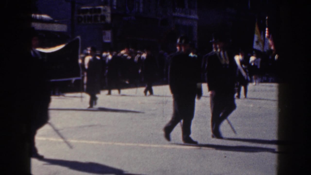 Men in dark suits march along a sunny city street while spectators stand behind them.