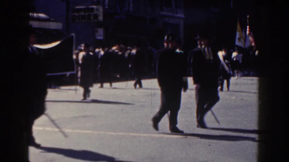 1962 New York City parade footage showing suited men crossing a sunlit street with flags and spectators in the background.