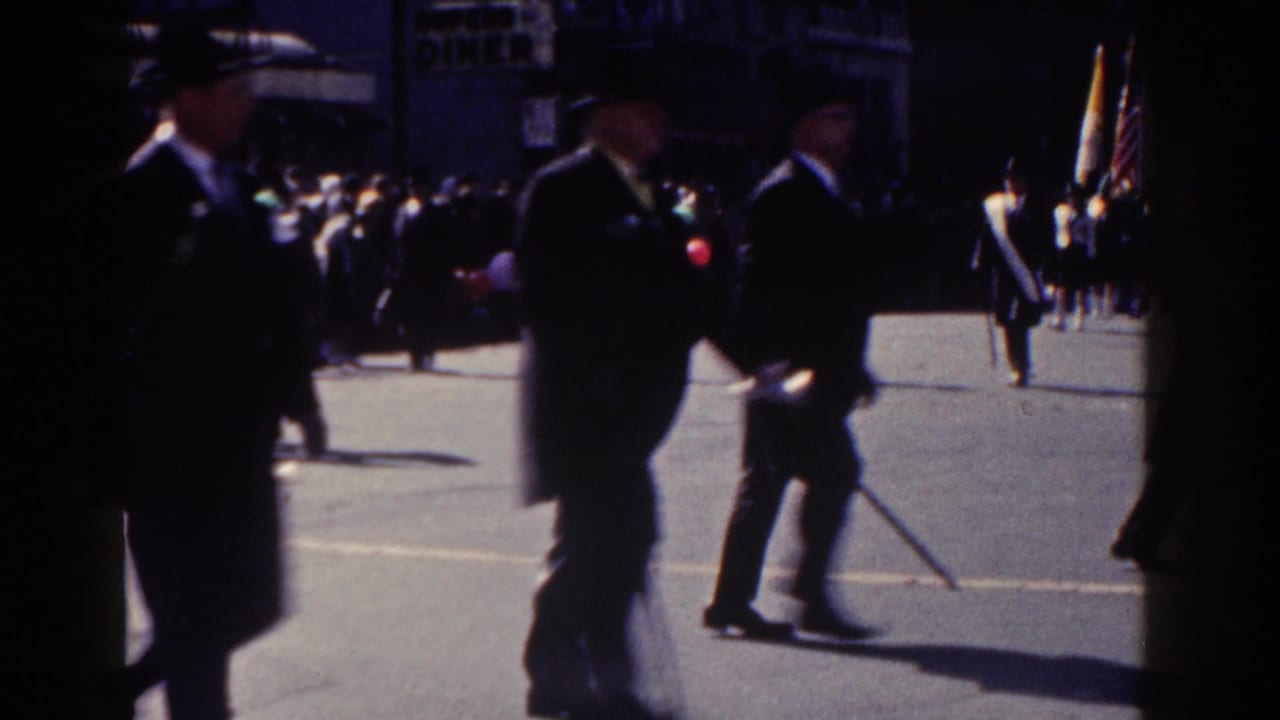 Suited parade marchers pass close to the 8mm camera in New York City, with spectators and flags visible across the street.