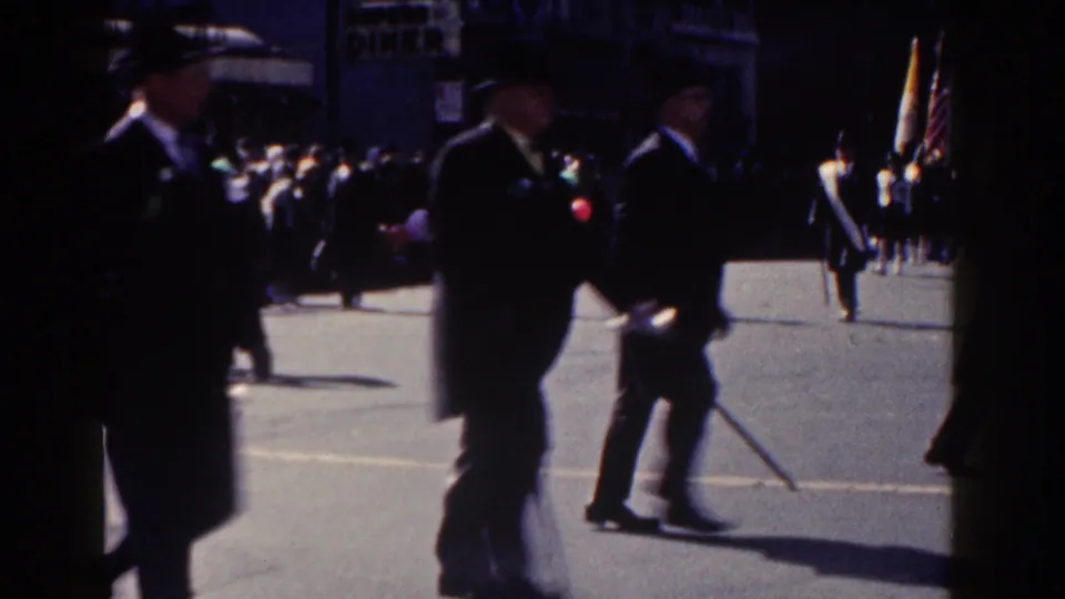 Suited parade marchers pass close to the 8mm camera in New York City, with spectators and flags visible across the street.