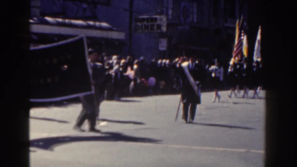 A dark banner carried through a 1962 New York City parade passes in front of a crowd and storefronts on a sunny street.