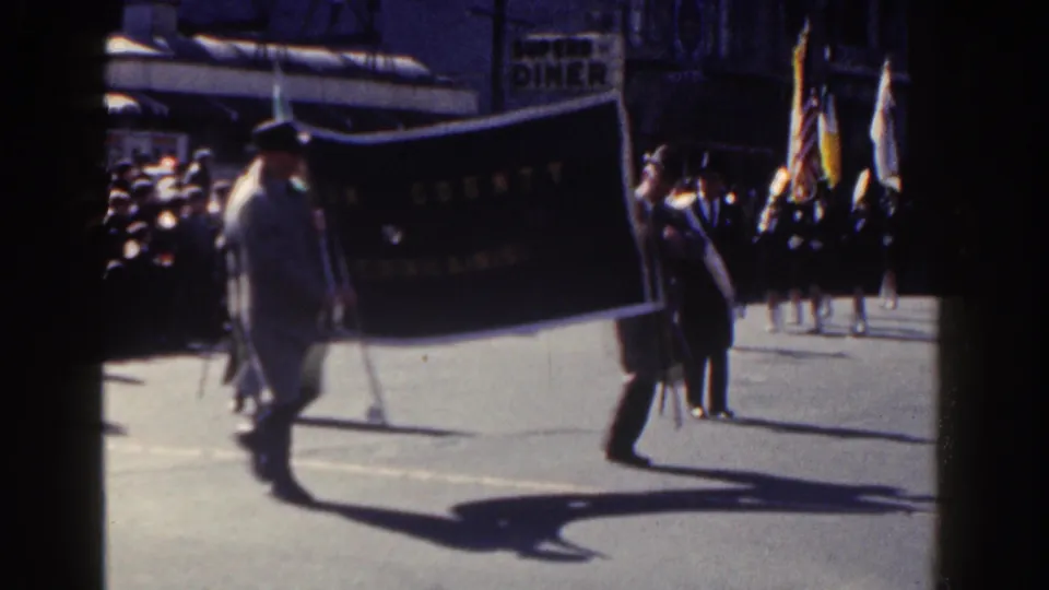 Late-frame view of a 1962 New York City parade with a dark banner, suited marcher, flags, and spectators along the street.
