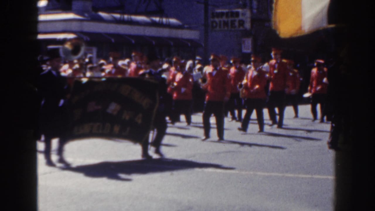 Marching band in red uniforms passes a city street crowd with a dark banner in front.