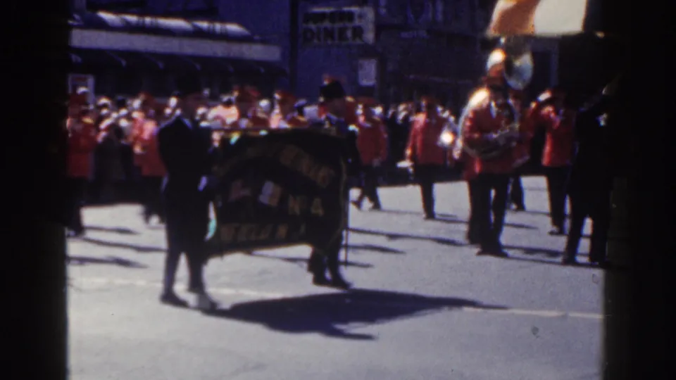 Street-level 1962 New York City parade view with red-uniformed musicians, a dark banner, spectators, and a diner sign in the background.
