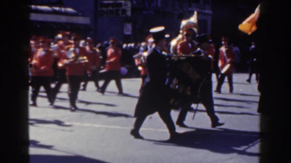 1962 New York City parade frame with dark-uniformed banner carriers crossing in front of red-uniformed band members and spectators.